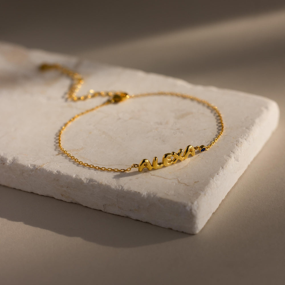 The gold “ALEXA” name bracelet with a sapphire birthstone is displayed in full on a light stone backdrop.