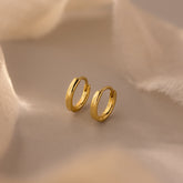 A pair of 7mm gold mini dainty huggie hoop earrings displayed on a soft beige fabric background with warm lighting.