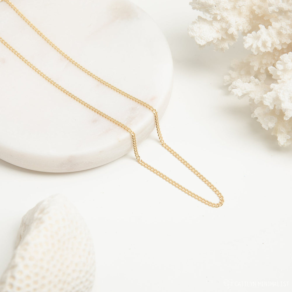 A gold curb chain necklace laid flat in a V shape on a round white marble tray, styled with white coral elements.