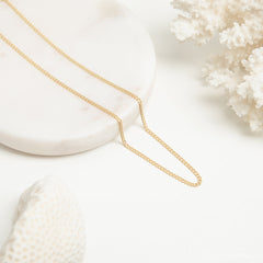 A gold curb chain necklace laid flat in a V shape on a round white marble tray, styled with white coral elements.
