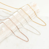 A variety of curb chain necklaces in gold, silver, and rose gold finishes displayed draped across a textured cream prop.