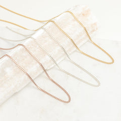 A variety of curb chain necklaces in gold, silver, and rose gold finishes displayed draped across a textured cream prop.