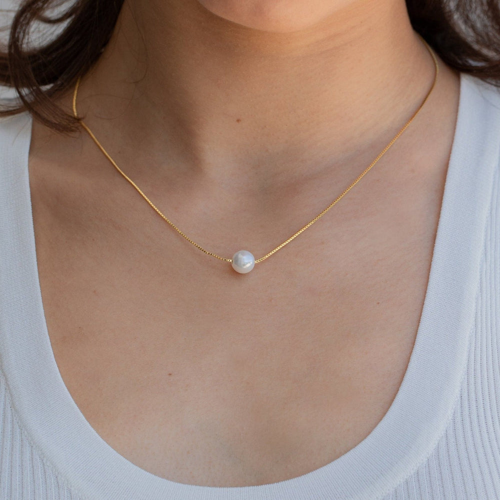 Woman wearing a minimalist gold chain necklace with a single round white pearl centered at the collarbone, styled with a white ribbed scoop-neck tank.