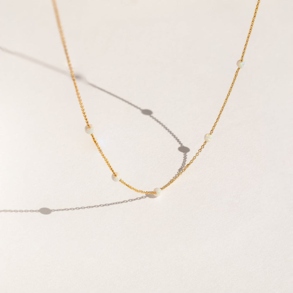 Delicate gold chain necklace with evenly spaced small opal beads, shown casting soft shadows on a light cream background.