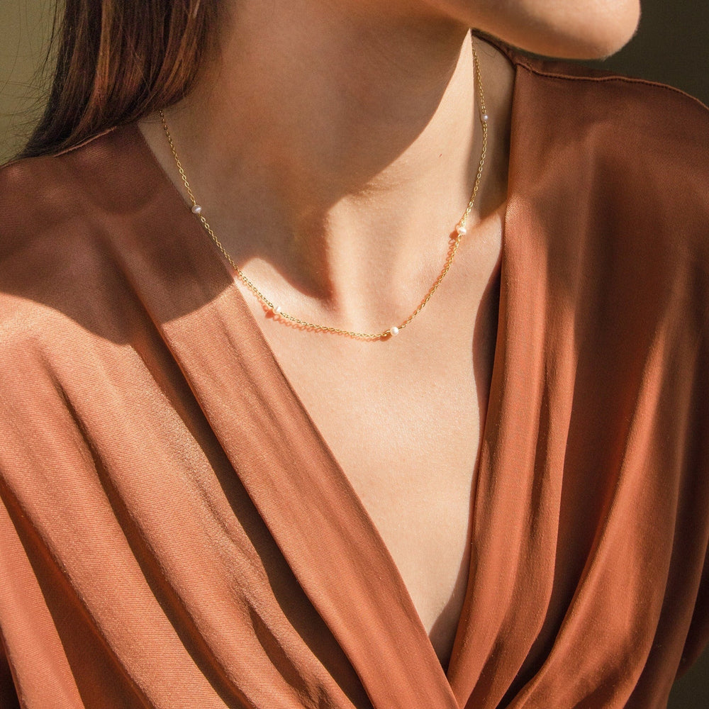 Delicate gold chain necklace with small evenly spaced pearls, worn by a woman in a deep V neck rust colored blouse.