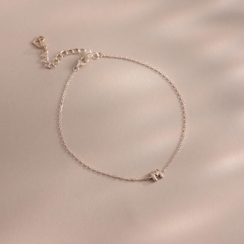 Close up of a dainty silver bubble letter M bracelet against a neutral background.