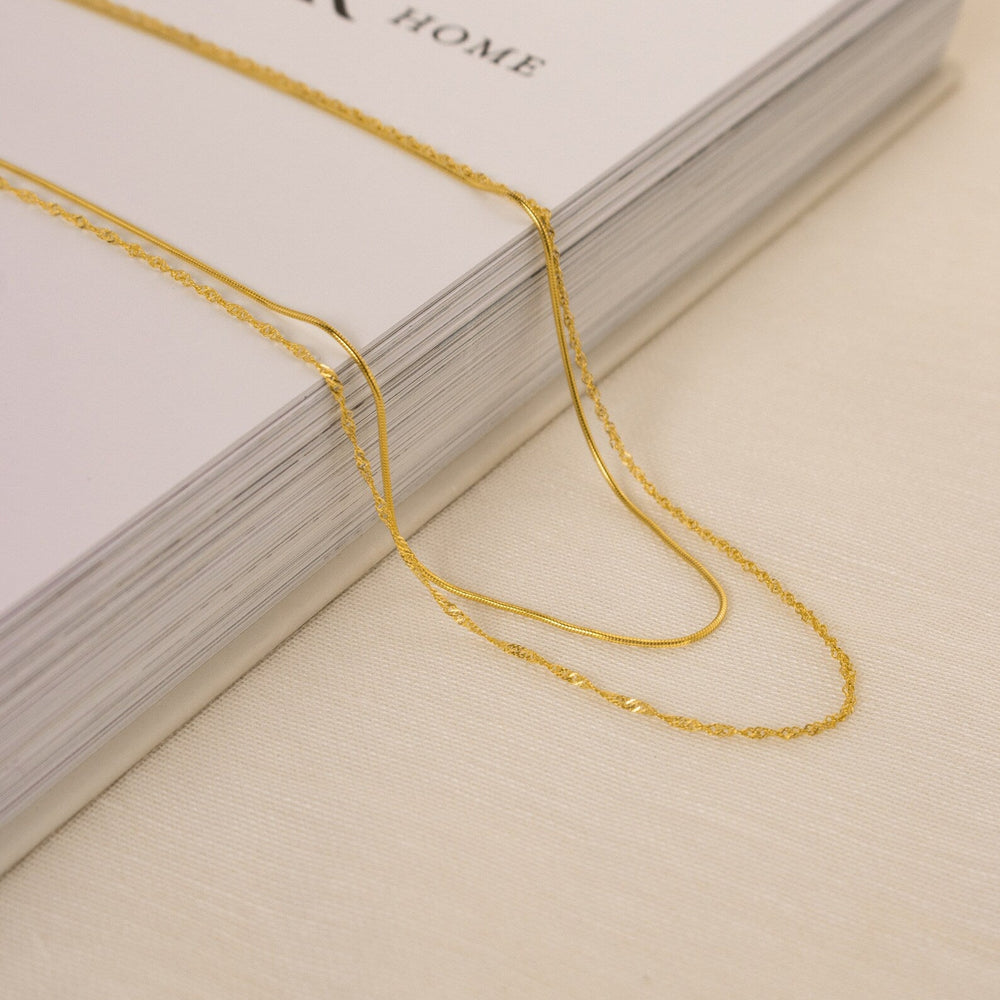 Two gold necklaces laid across a thick hardcover book: one features a sleek snake chain and the other a delicate faceted bead-style chain, both resting on a cream fabric surface.
