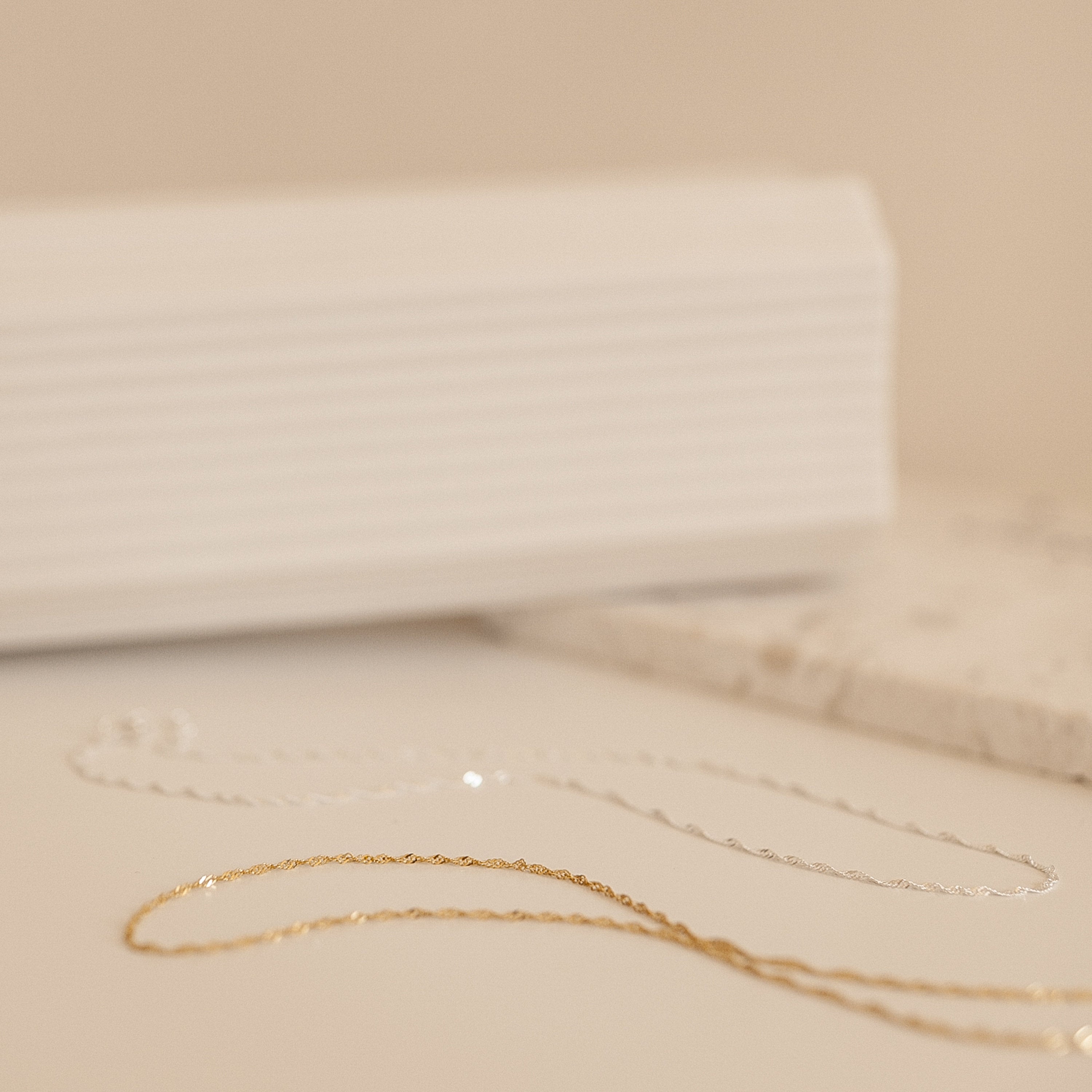 A soft-focus image of gold and silver Singapore Twist necklaces arranged flatly on a neutral-toned surface.